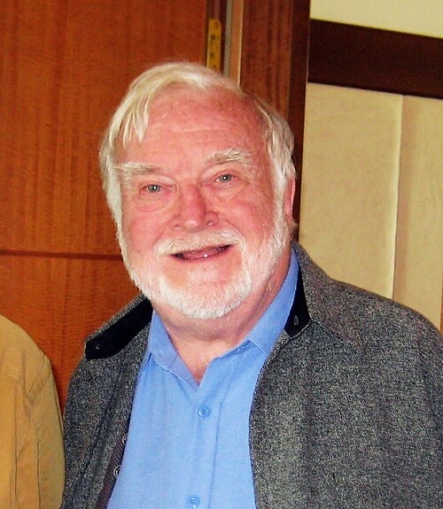 Mihaly Csikszentmihalyi in 2010, smiling, white hair and beard, wearing a grey jacket over a blue shirt.