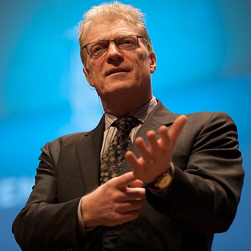 Ken Robinson speaking on stage against a blue backdrop, mid-gesture with one hand raised, wearing glasses and a dark suit.