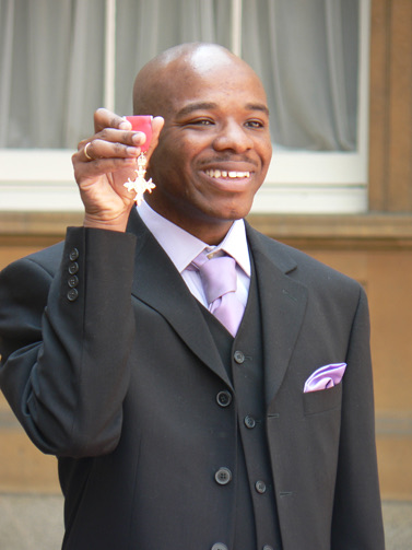 Stephen Wiltshire smiling outside Buckingham Palace in a dark overcoat and lilac tie, holding his MBE medal up in his right hand.